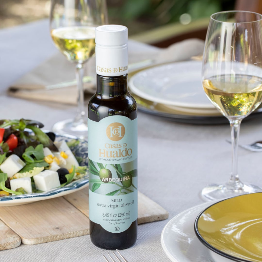 A bottle of CASAS DE HUALDO Extra Virgin Olive Oil Arbequina graces the table with a vibrant salad and two glasses of white wine, setting the scene for a charming outdoor dining experience, complete with elegant plates and cutlery.