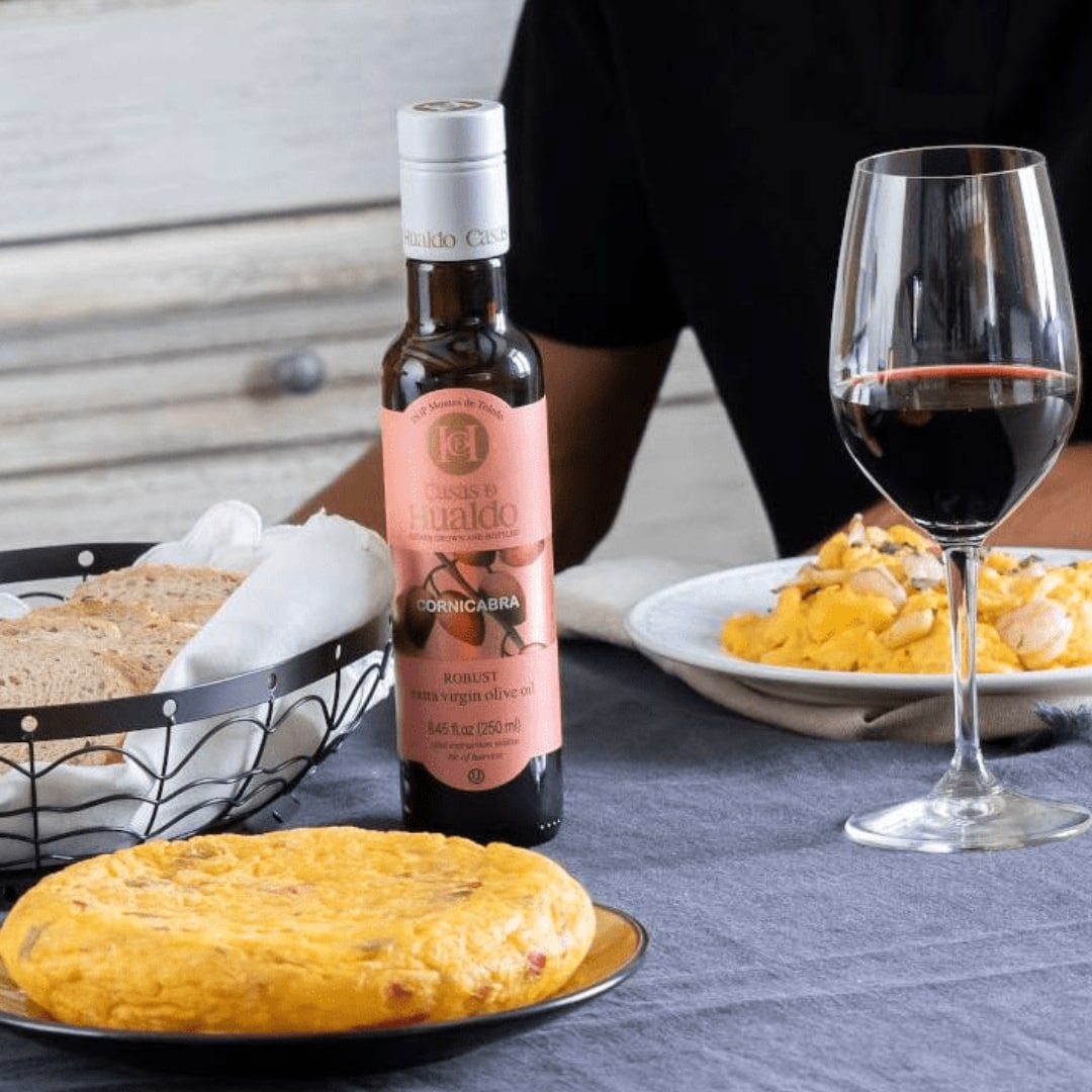 A table featuring a bottle of Casas de Hualdo Extra Virgin Olive Oil Cornicabra, a glass of red wine, a basket with sliced bread, and plates of Spanish tortilla and scrambled eggs; a seated person is partially visible.