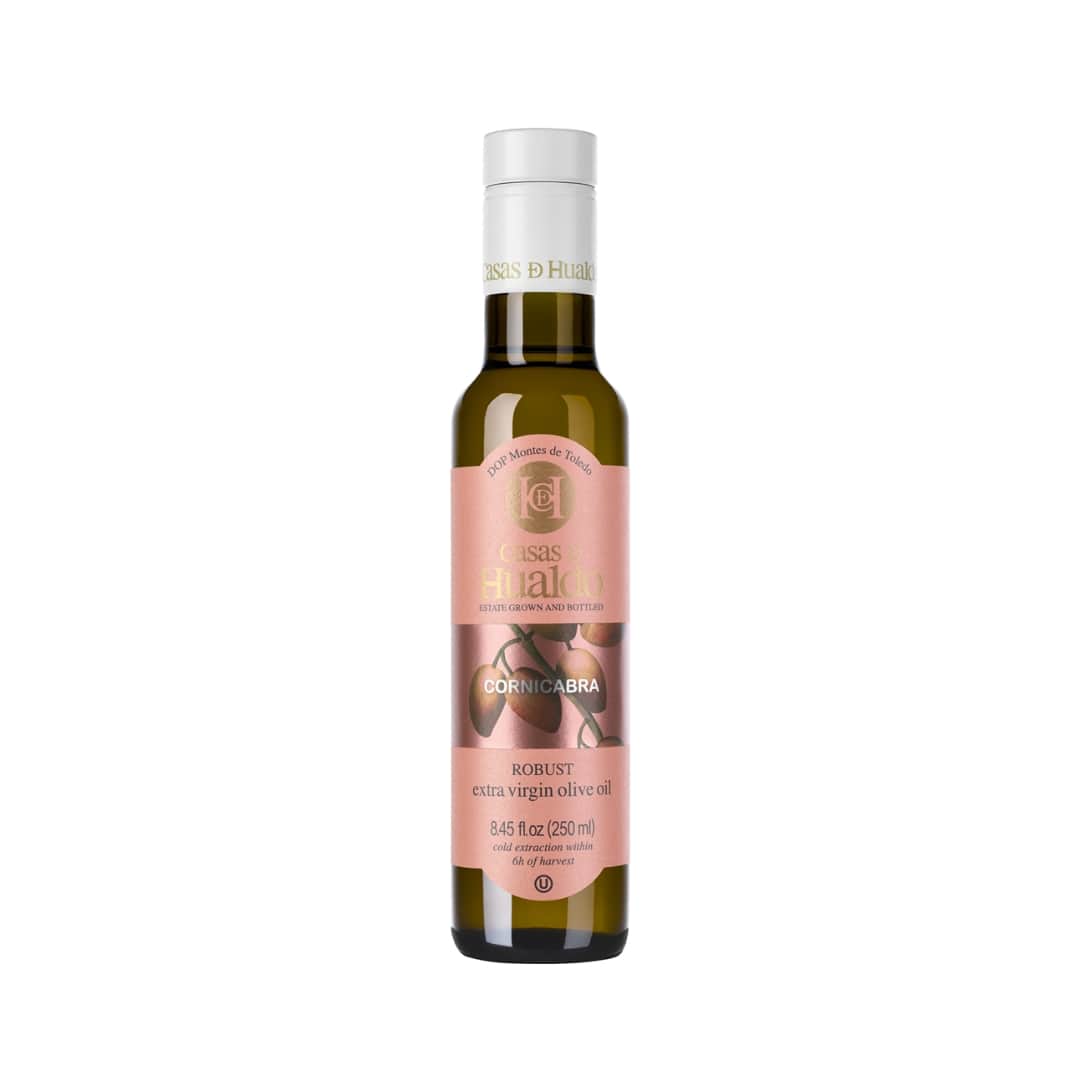 A 250 ml bottle of Casas de Hualdo Extra Virgin Olive Oil Cornicabra boasts its robust Spanish heritage with a pink label accented in gold and brown, featuring an olive image.