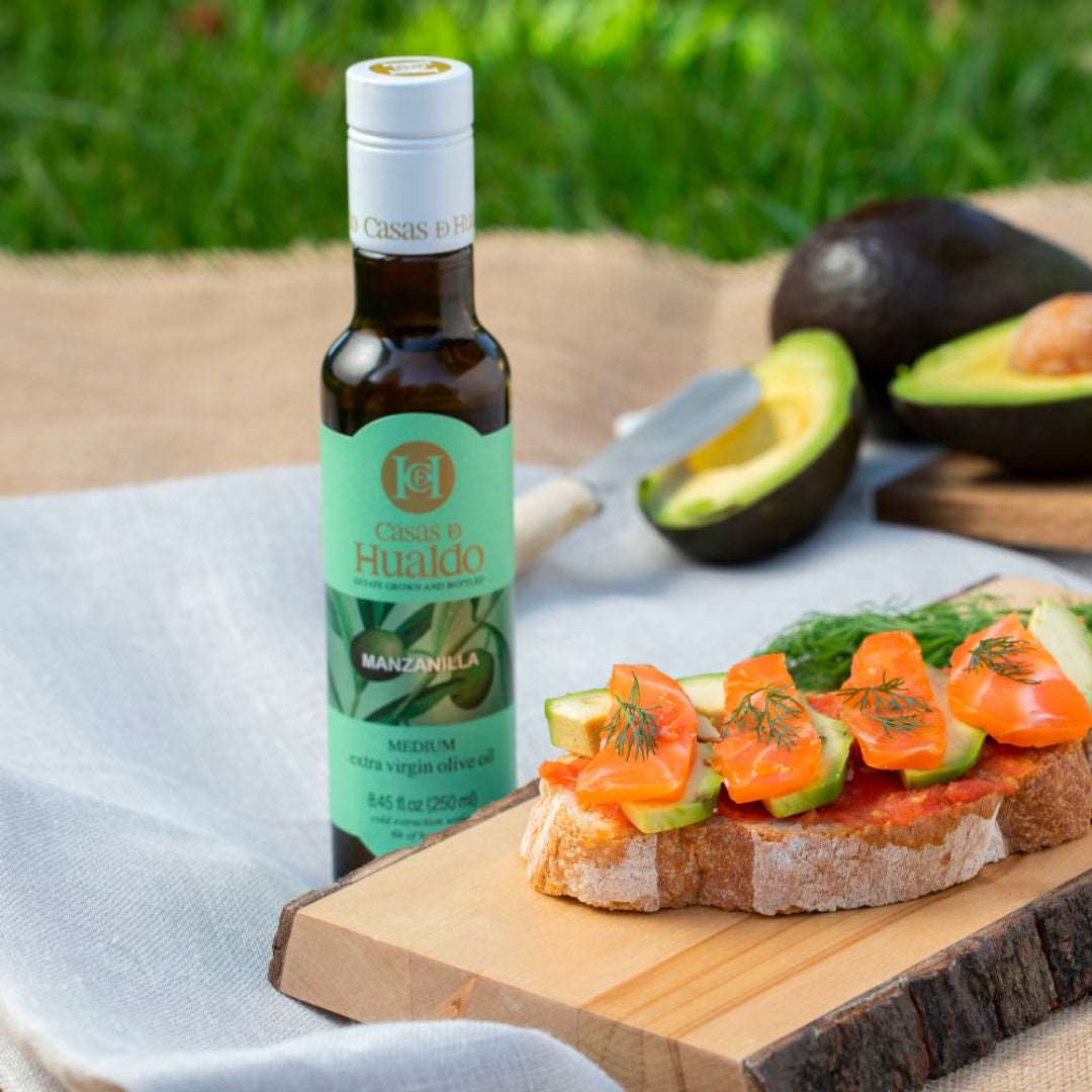 A bottle of CASAS DE HUALDO Extra Virgin Olive Oil Manzanilla sits beside a wooden board with bread, salmon, cucumber, and dill. In the backdrop, sliced and whole avocados rest on grass.