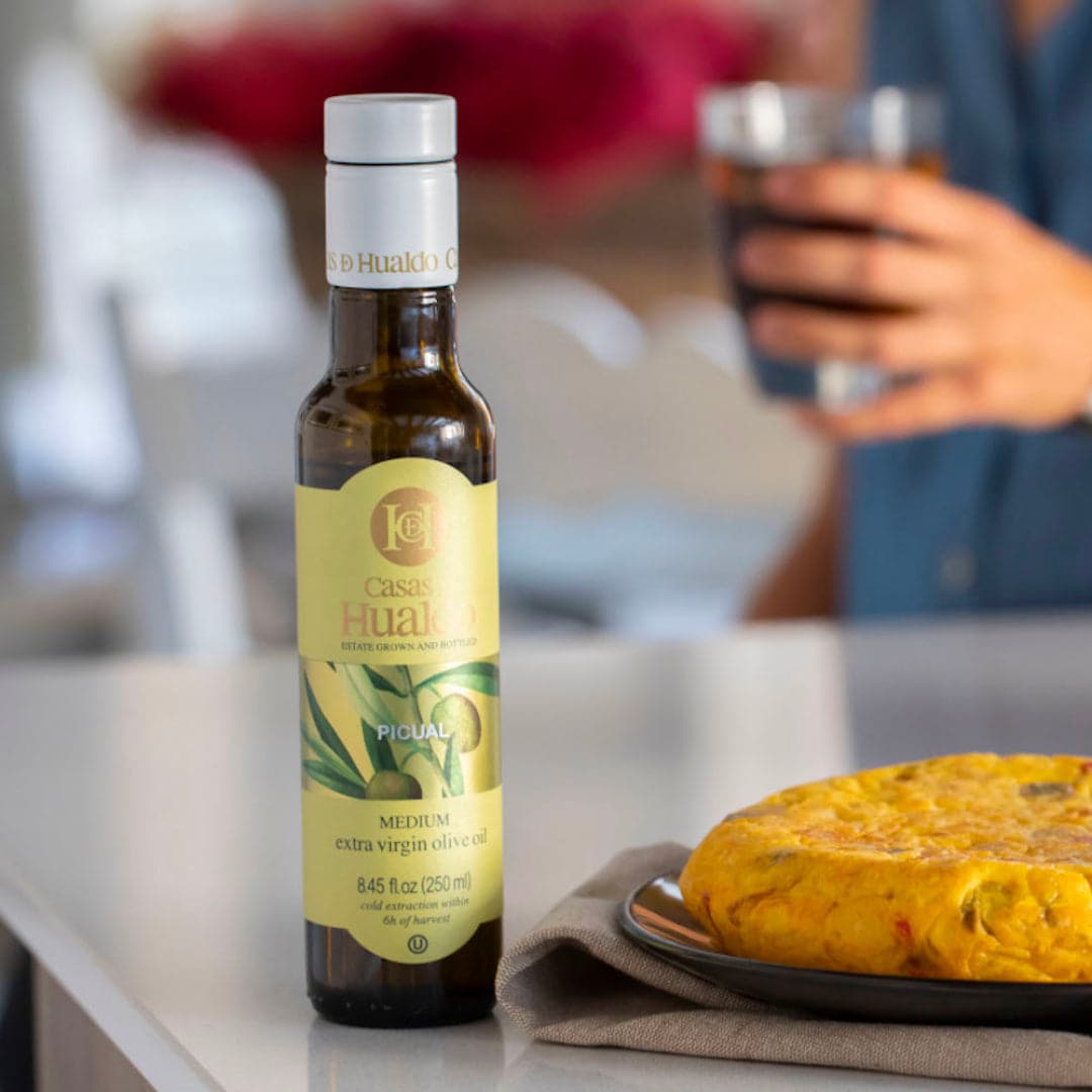 A bottle of CASAS DE HUALDO Extra Virgin Olive Oil Picual sits beside a Spanish tortilla, enriching the scene with its flavor. In the background, someone holds a glass of dark beverage, completing this inviting meal setting with genuine Spanish olive oil ambiance.