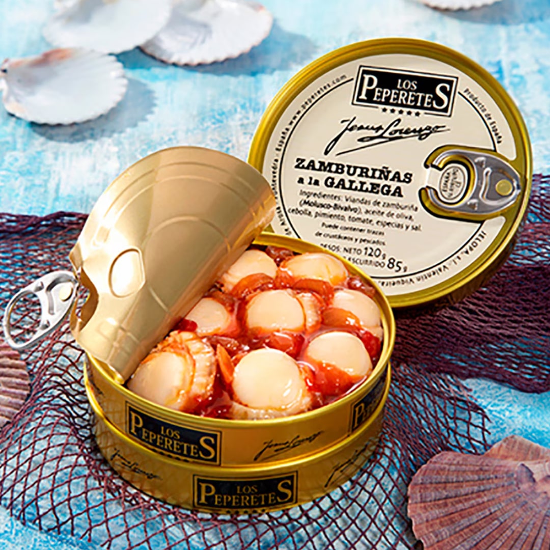 Open a can of LOS PEPERETES Galician-Style Scallops on a blue surface; its lid is slightly lifted, revealing tender scallops in sauce. Scallop shells and a decorative net enhance the coastal charm in the background.