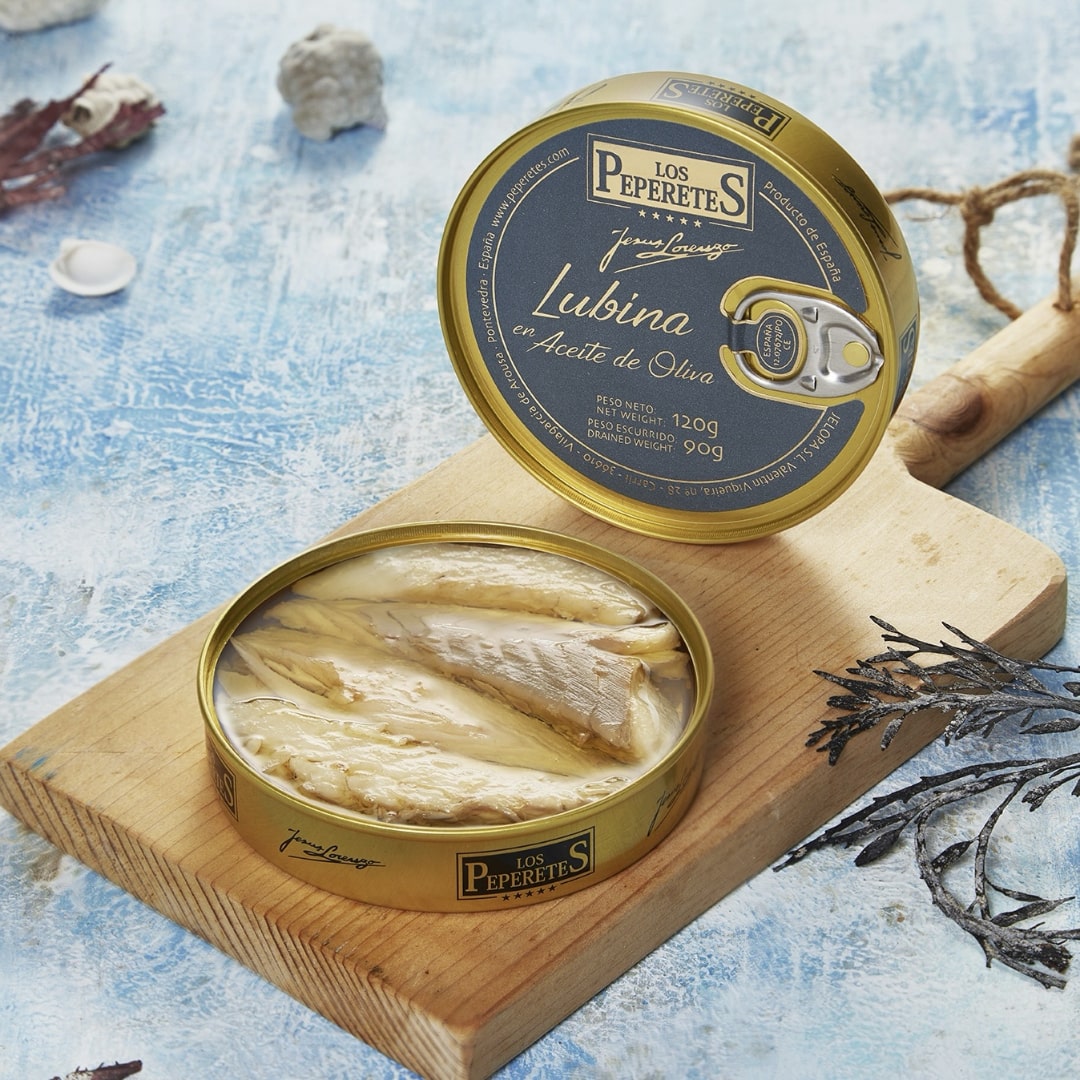 An open circular tin of LOS PEPERETES Sea Bass Fillets in Olive Oil reveals gourmet sea bass on a wooden board against a blue textured background, surrounded by decorative items.